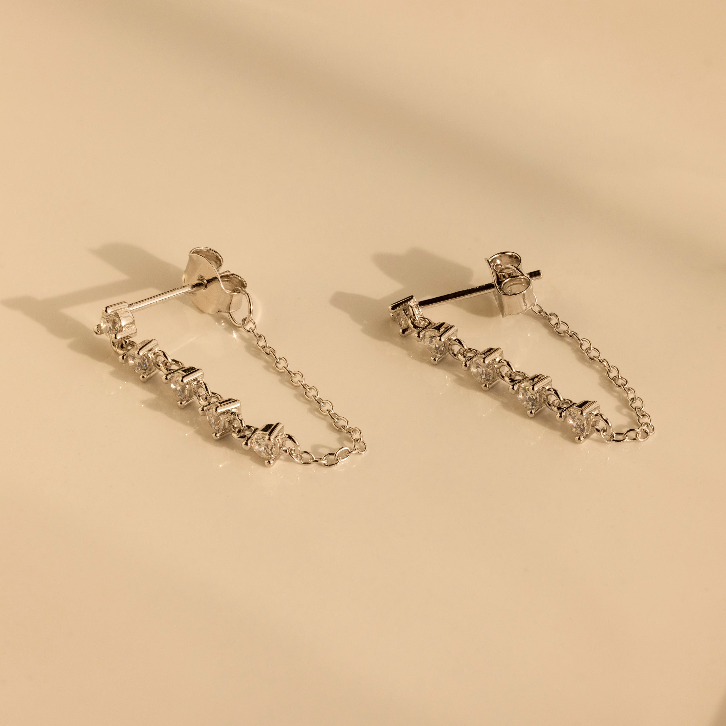 Diamond Chain Drop Earrings in Silver feature delicate chains and small crystals, elegantly displayed on a smooth beige surface.
