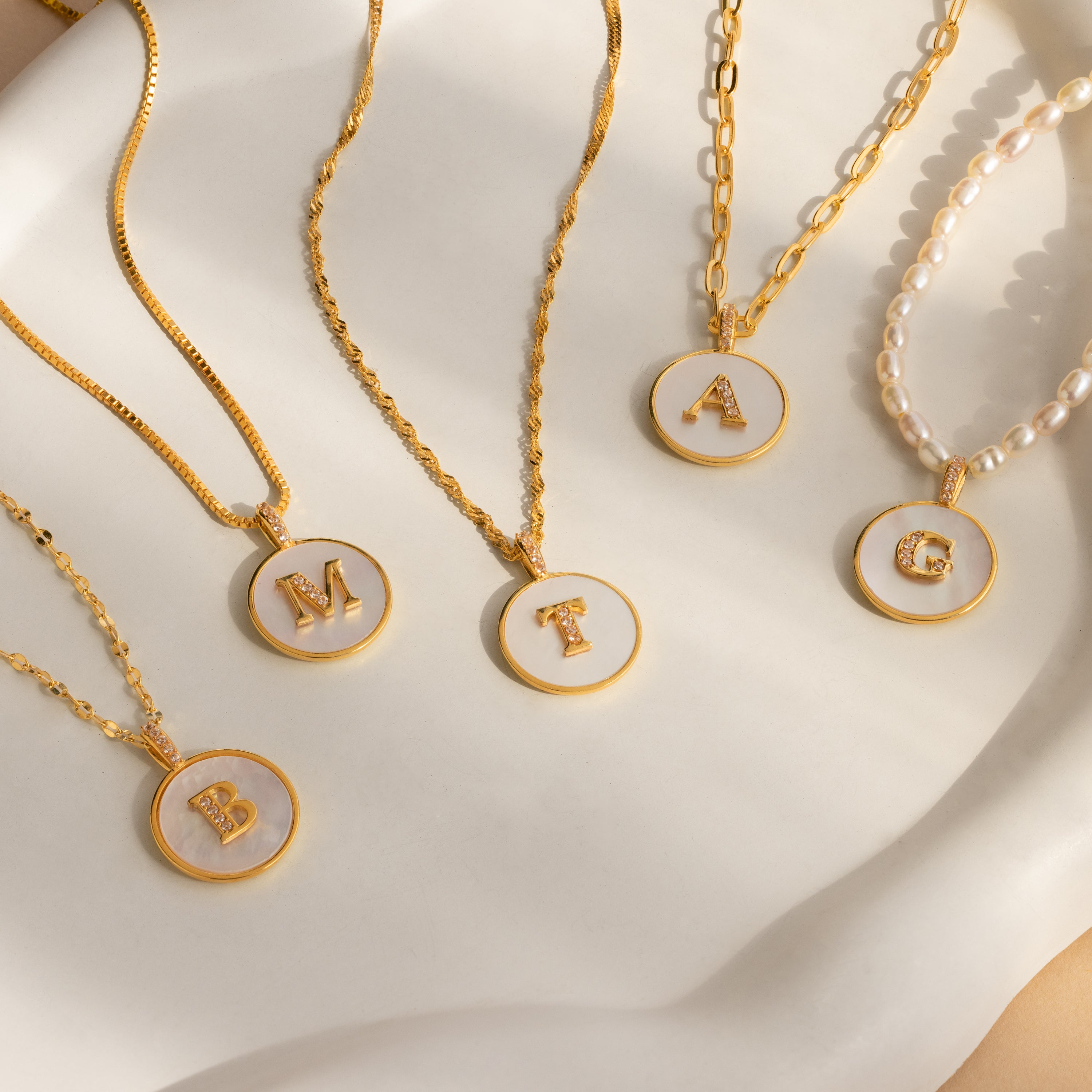 Five Pearl Initial Disc Necklaces with round pendants featuring the letters B, M, T, A, and G on a white surface—a beautiful personalized or custom initial gift for any occasion.