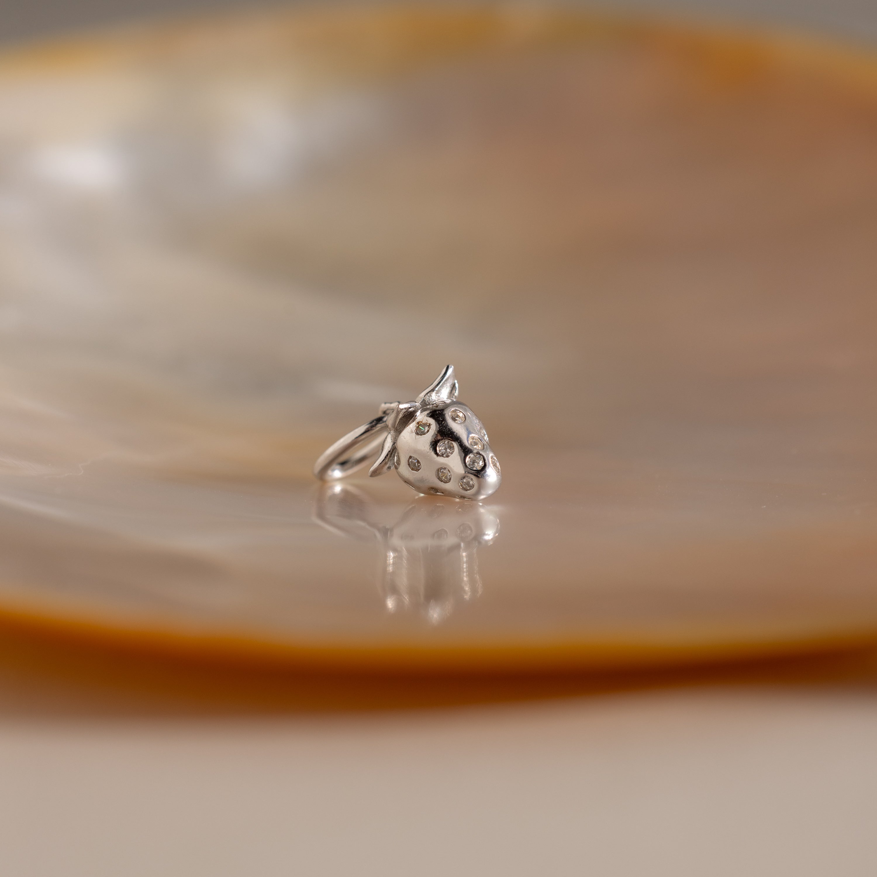 Silver strawberry charm with small crystal accents, resting on a glossy, shell-like surface.