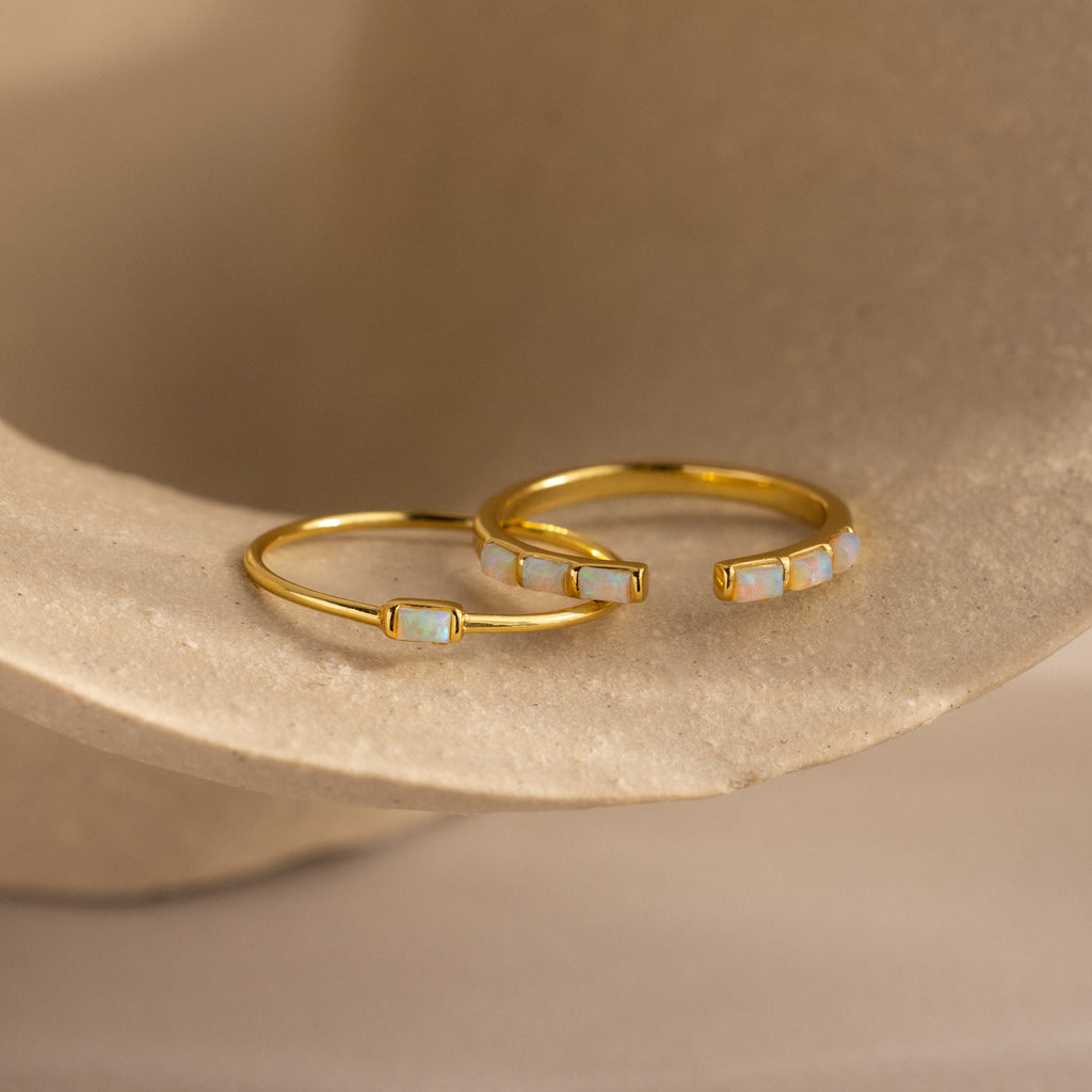 Two dainty gold rings with baguette-cut opals, one featuring a single opal and the other adorned with five opals in a row.