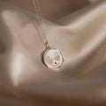 The Baby Hand & Footprint Necklace in Sterling Silver features a pendant with two tiny handprints and the name Reagan, beautifully displayed on silky beige fabric—an ideal piece of personalized baby jewelry.