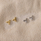 Two pairs of small, metallic Shark Tooth Flatback Studs—one gold, one silver—resting on a sandy surface make the perfect beach accessory.