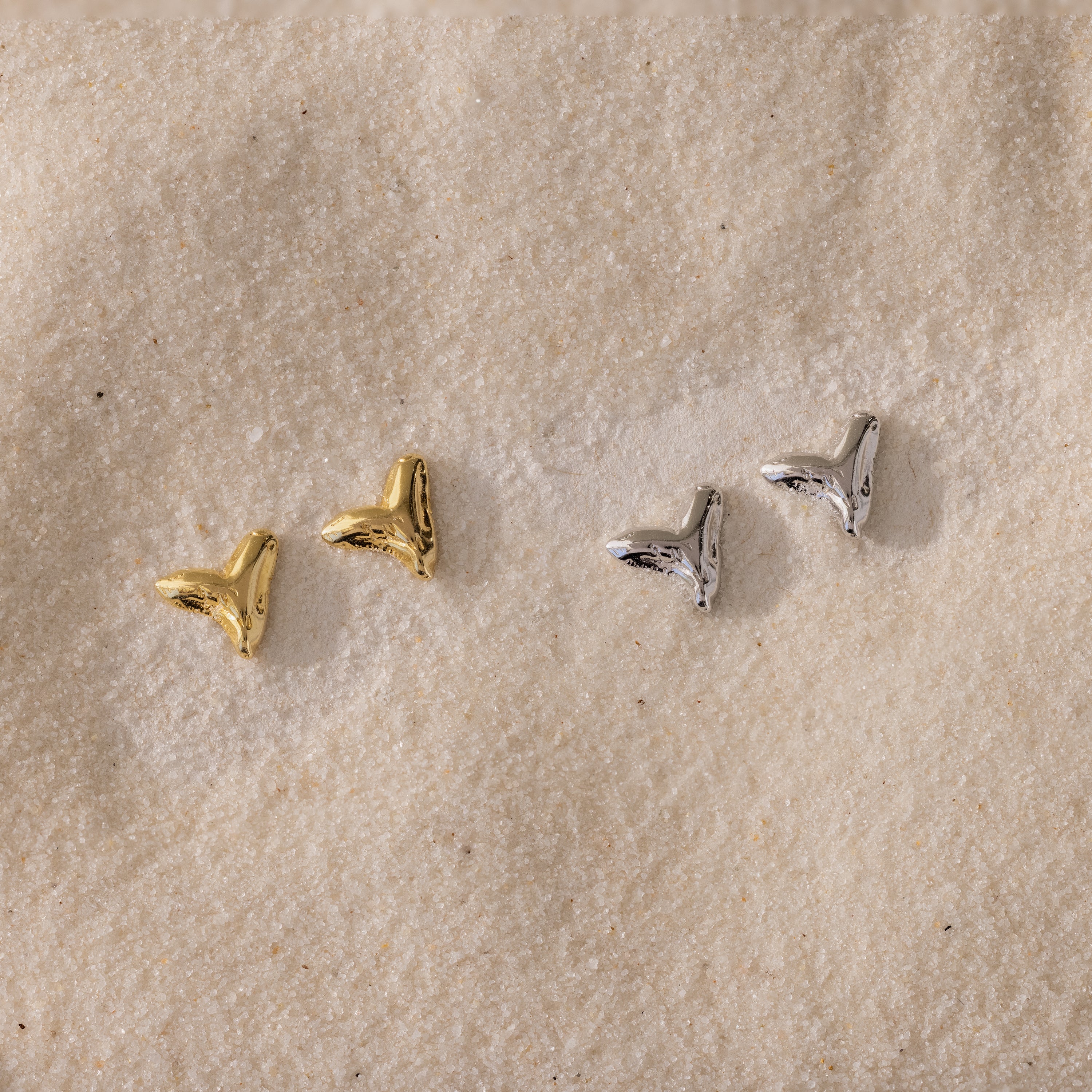Two pairs of small, metallic Shark Tooth Flatback Studs—one gold, one silver—resting on a sandy surface make the perfect beach accessory.