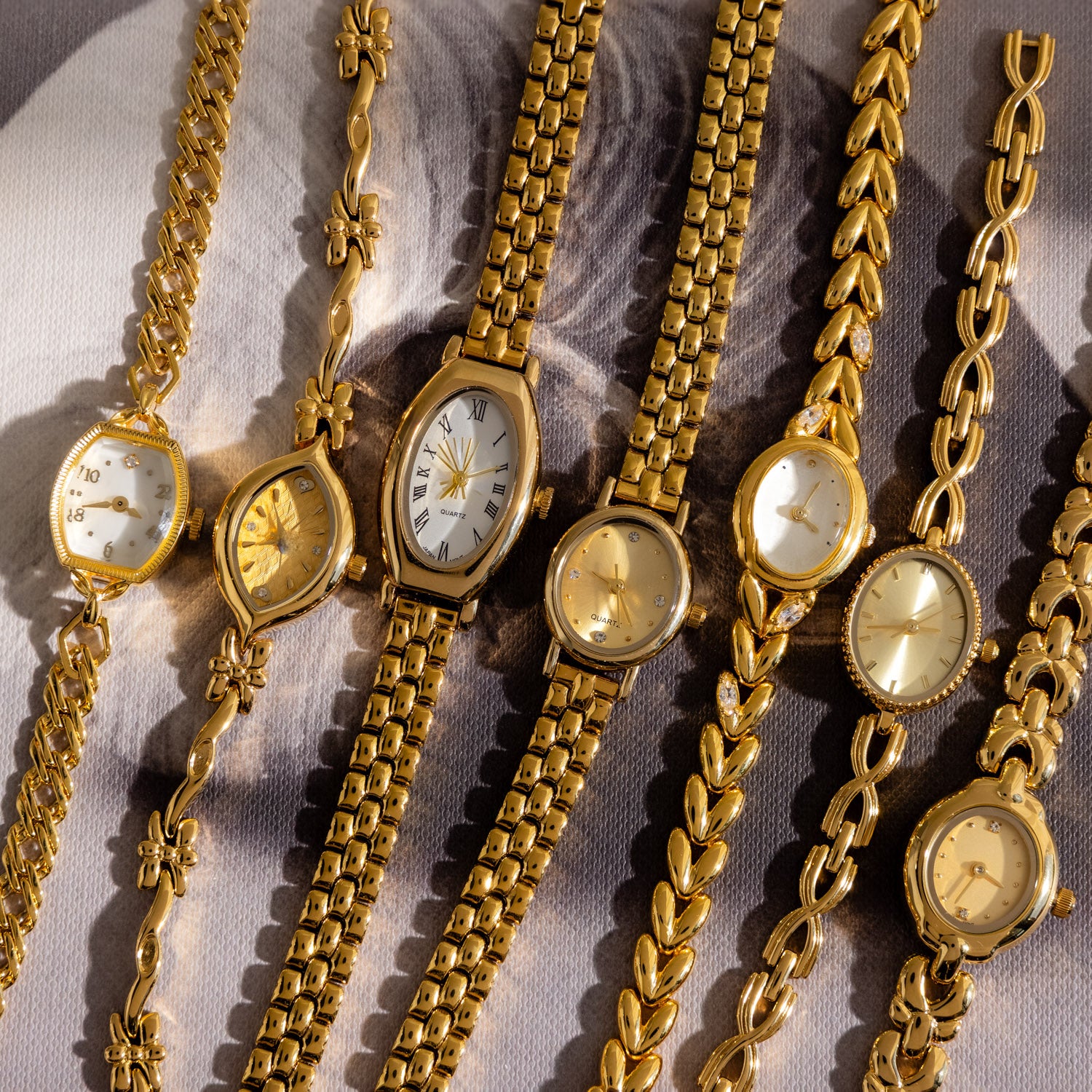 Seven Heirloom Chain Link Timepieces, featuring various designs like diamond accents and mother of pearl dials, are arranged side by side on a patterned background.