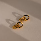 Two pairs of Twisted Link Earrings in 18K gold, their interlocking forms casting soft shadows on a beige surface—highlighting elegant jewelry and modern design.