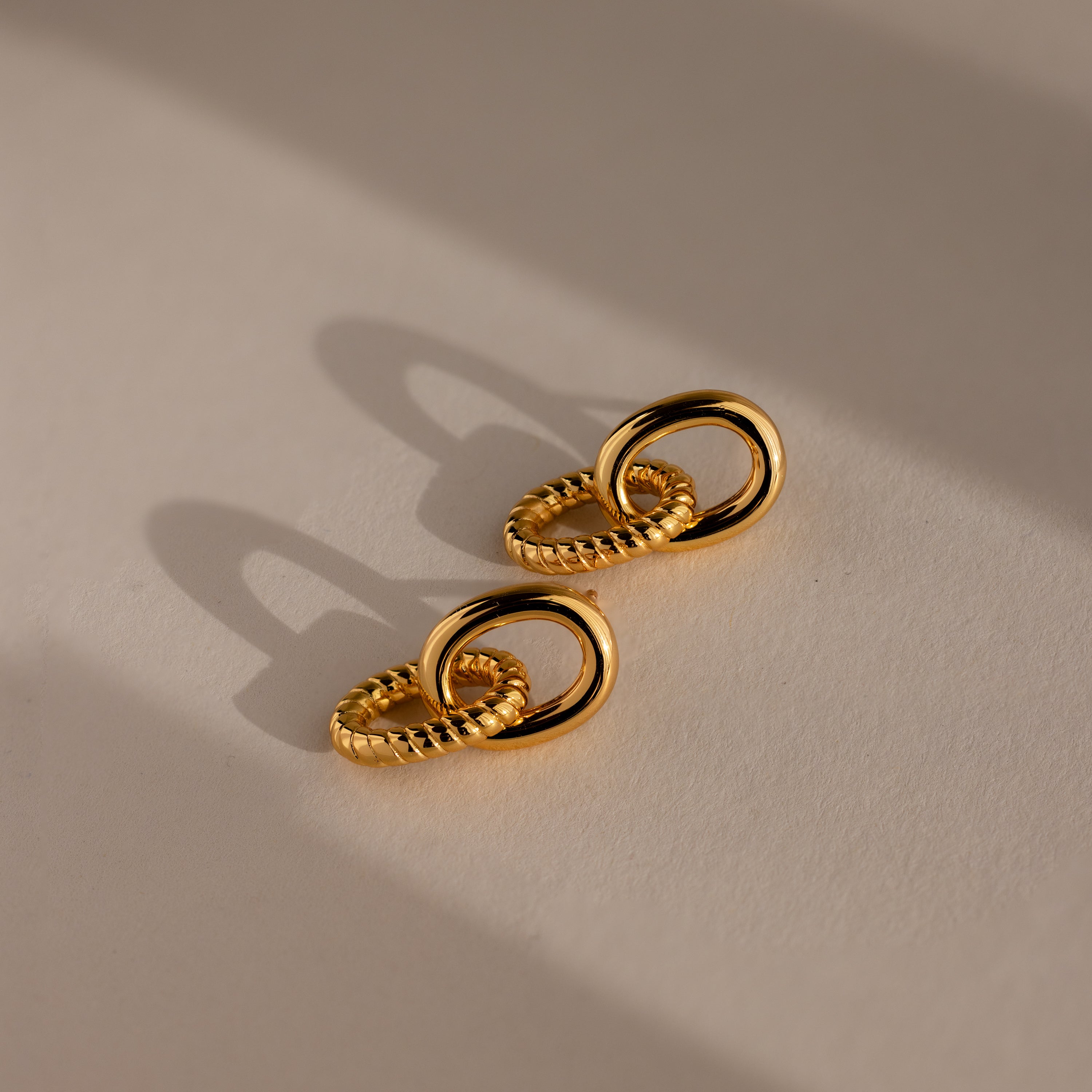 Two pairs of Twisted Link Earrings in 18K gold, their interlocking forms casting soft shadows on a beige surface—highlighting elegant jewelry and modern design.