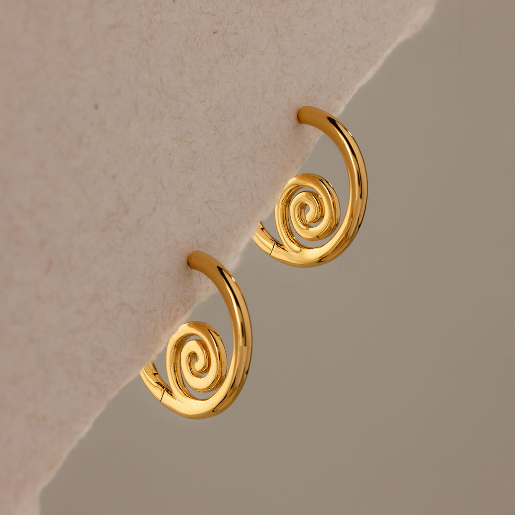 Spiral Swirl Hoops in 18K Gold rest on a soft beige fabric—ideal beachy earrings to elevate your next boho vacation jewelry collection.