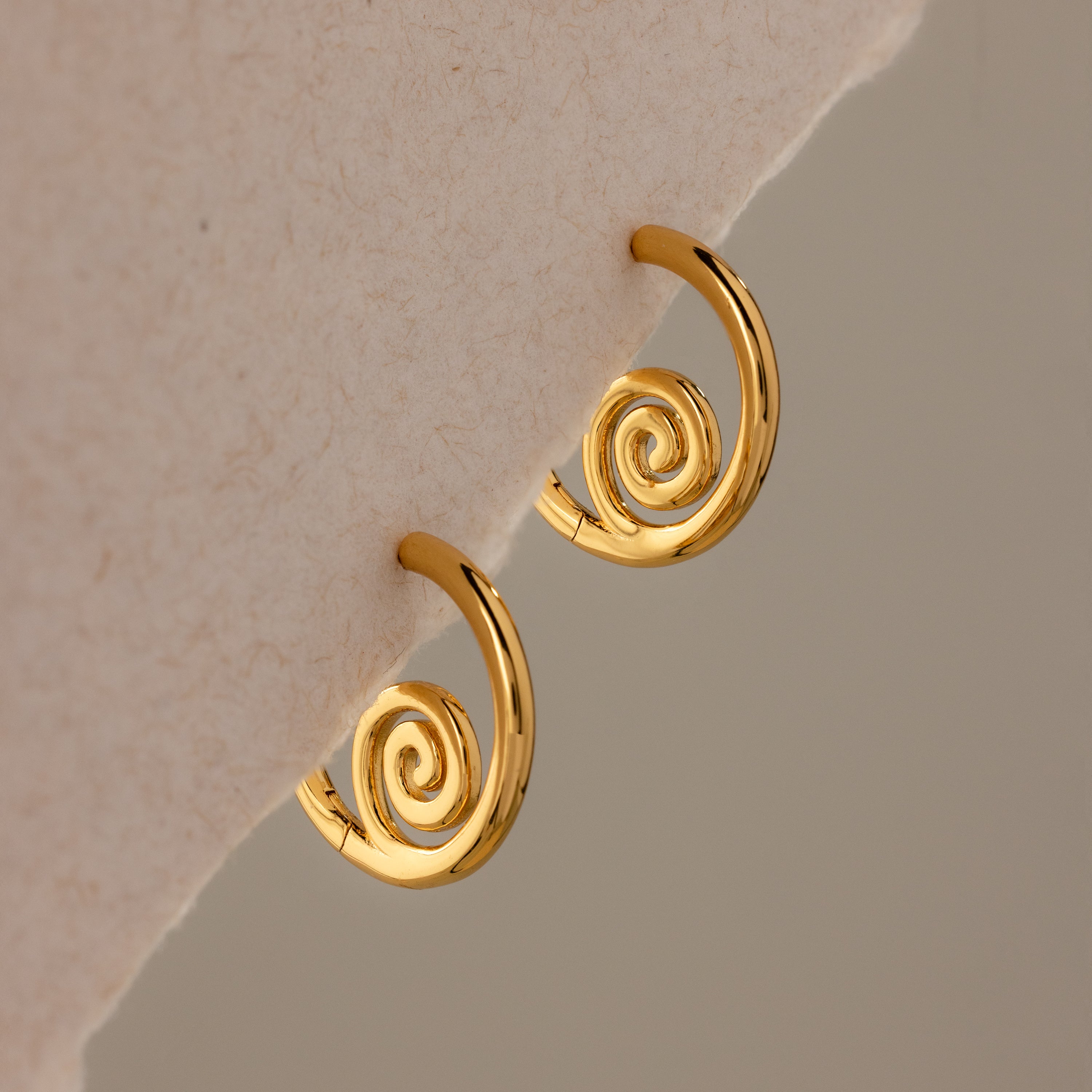 Spiral Swirl Hoops in 18K Gold rest on a soft beige fabric—ideal beachy earrings to elevate your next boho vacation jewelry collection.