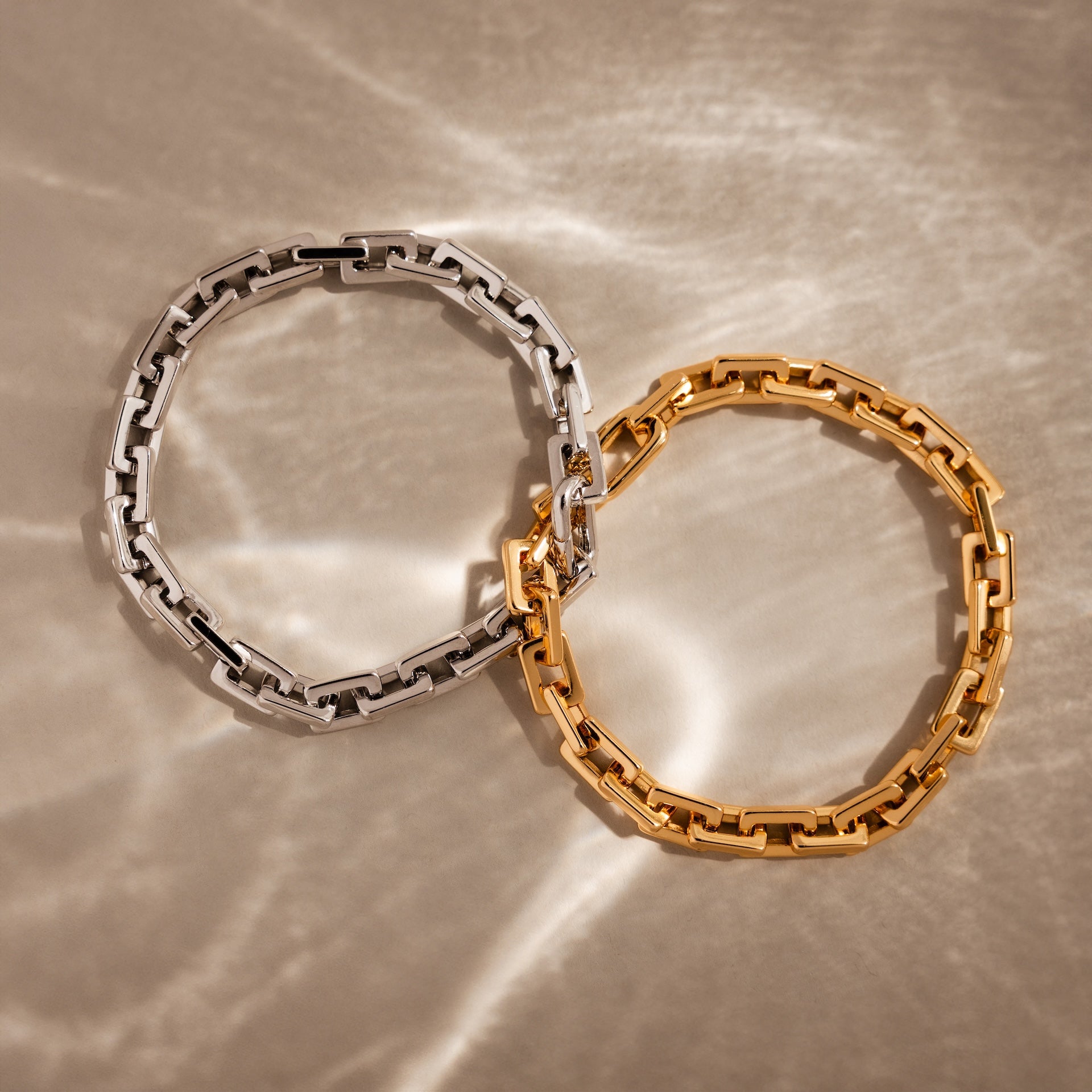 The Eternal Link Bracelet, in both silver and gold, arranged on a softly textured beige surface with light reflections—perfect modern essentials.