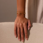 A hand with nude nails rests on a beige surface, showcasing three delicate gold chain bracelets, including the Eternal Link Bracelet in 18K Gold with a subtle geometric design.