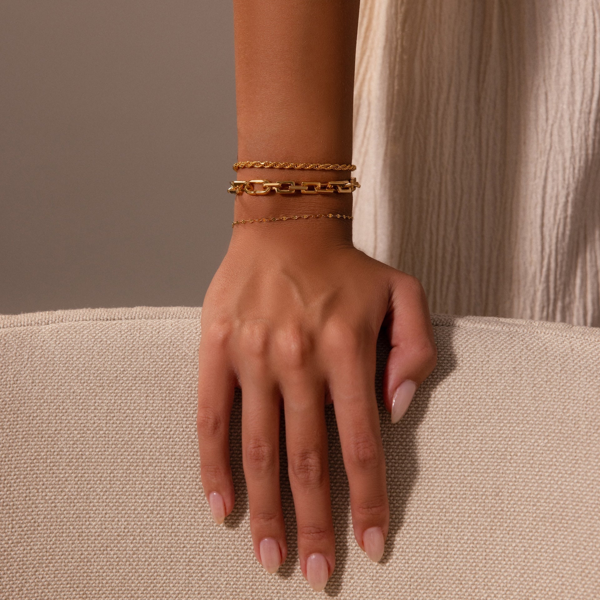 A hand with nude nails rests on a beige surface, showcasing three delicate gold chain bracelets, including the Eternal Link Bracelet in 18K Gold with a subtle geometric design.
