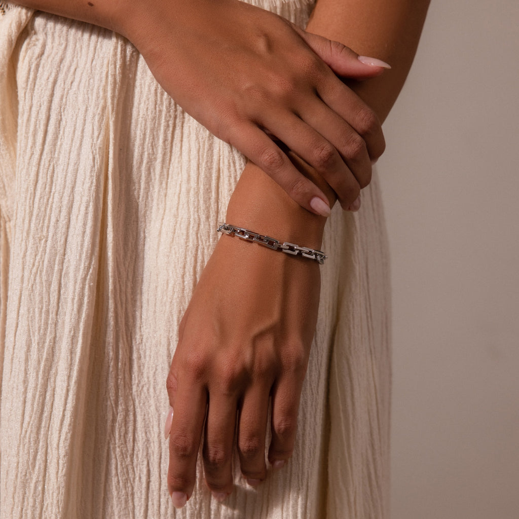 A person wearing the Eternal Link Bracelet, a modern essential with a geometric design, gently crosses their hands while dressed in a cream pleated skirt.