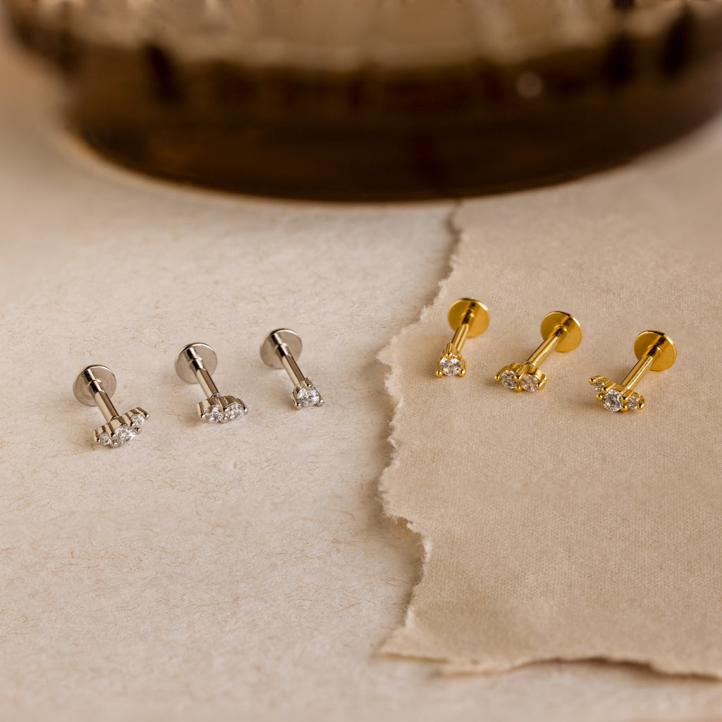 Two pairs of silver and two pairs of gold Tiny Diamond Flatback Studs with clear stones rest on a beige textured surface.