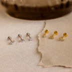Two pairs of silver and two pairs of gold Tiny Diamond Flatback Studs with clear stones rest on a beige textured surface.