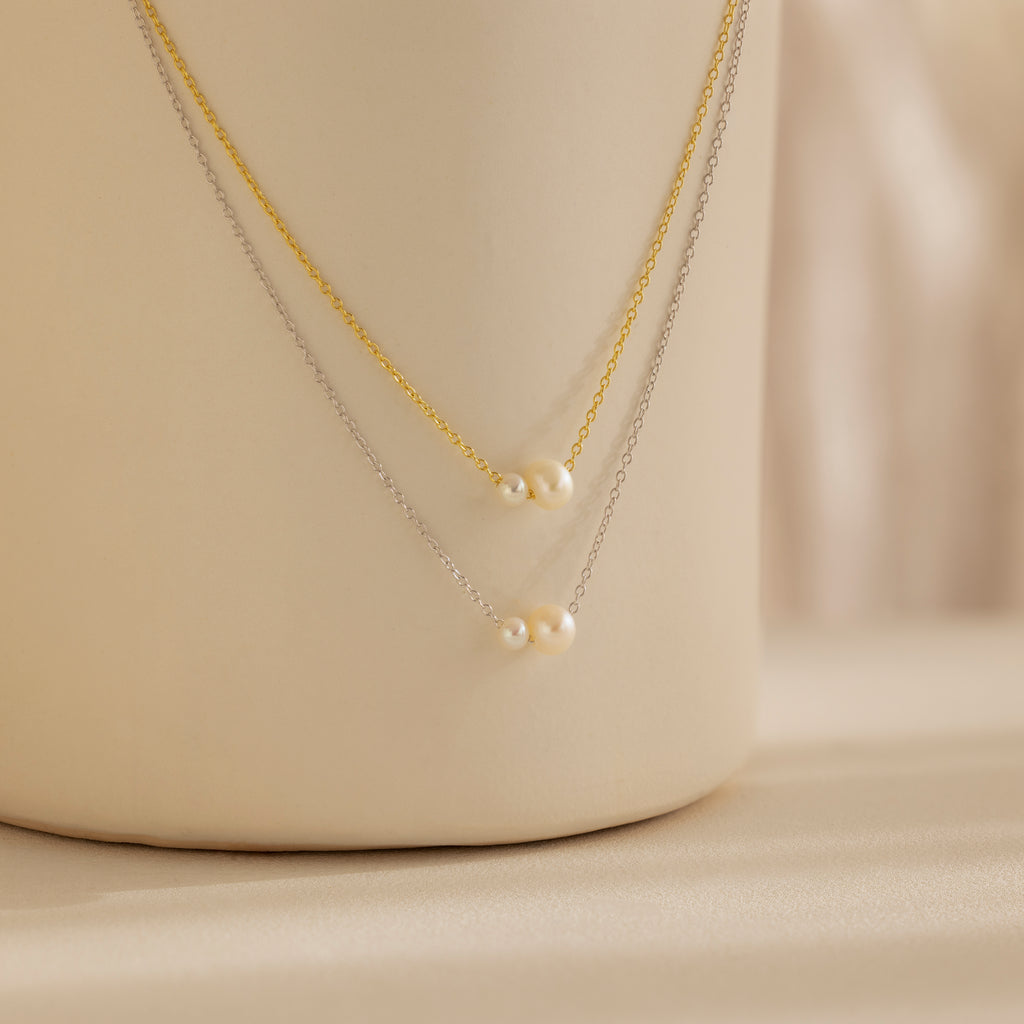 The Dainty Duo Pearl Necklace features gold and silver chains, each accented with small pearls, elegantly displayed on a cream surface—ideal as wedding jewelry or a chic accessory for any occasion.