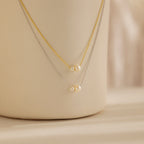 The Dainty Duo Pearl Necklace features gold and silver chains, each accented with small pearls, elegantly displayed on a cream surface—ideal as wedding jewelry or a chic accessory for any occasion.