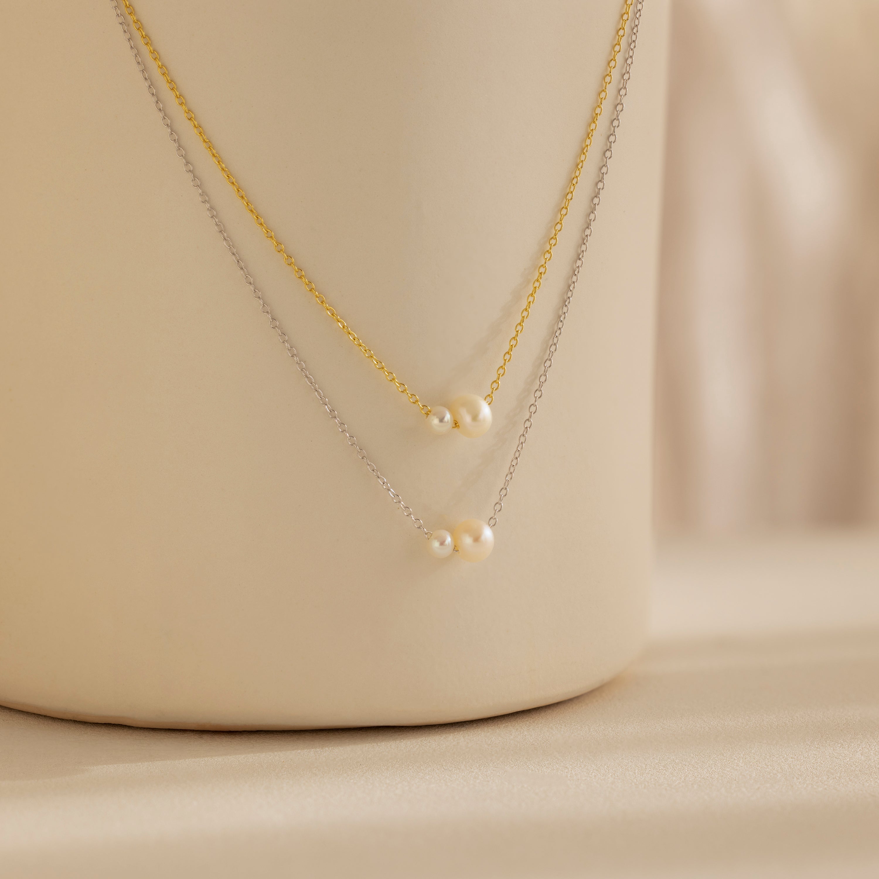The Dainty Duo Pearl Necklace features gold and silver chains, each accented with small pearls, elegantly displayed on a cream surface—ideal as wedding jewelry or a chic accessory for any occasion.