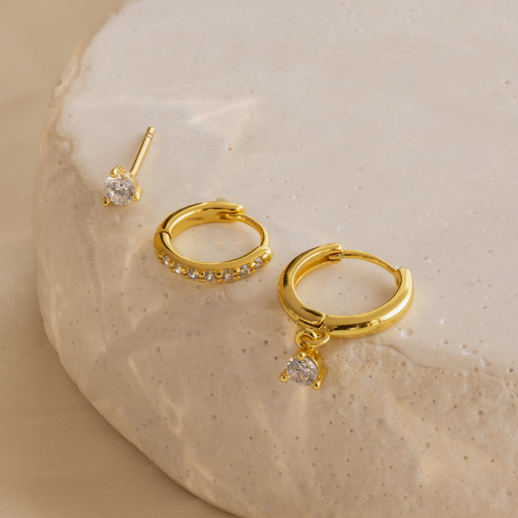 The Classic Diamond Earrings Set in 18K Gold features three pairs with clear gemstones, displayed on a textured beige surface—perfect for adding elegance to any bridal look.