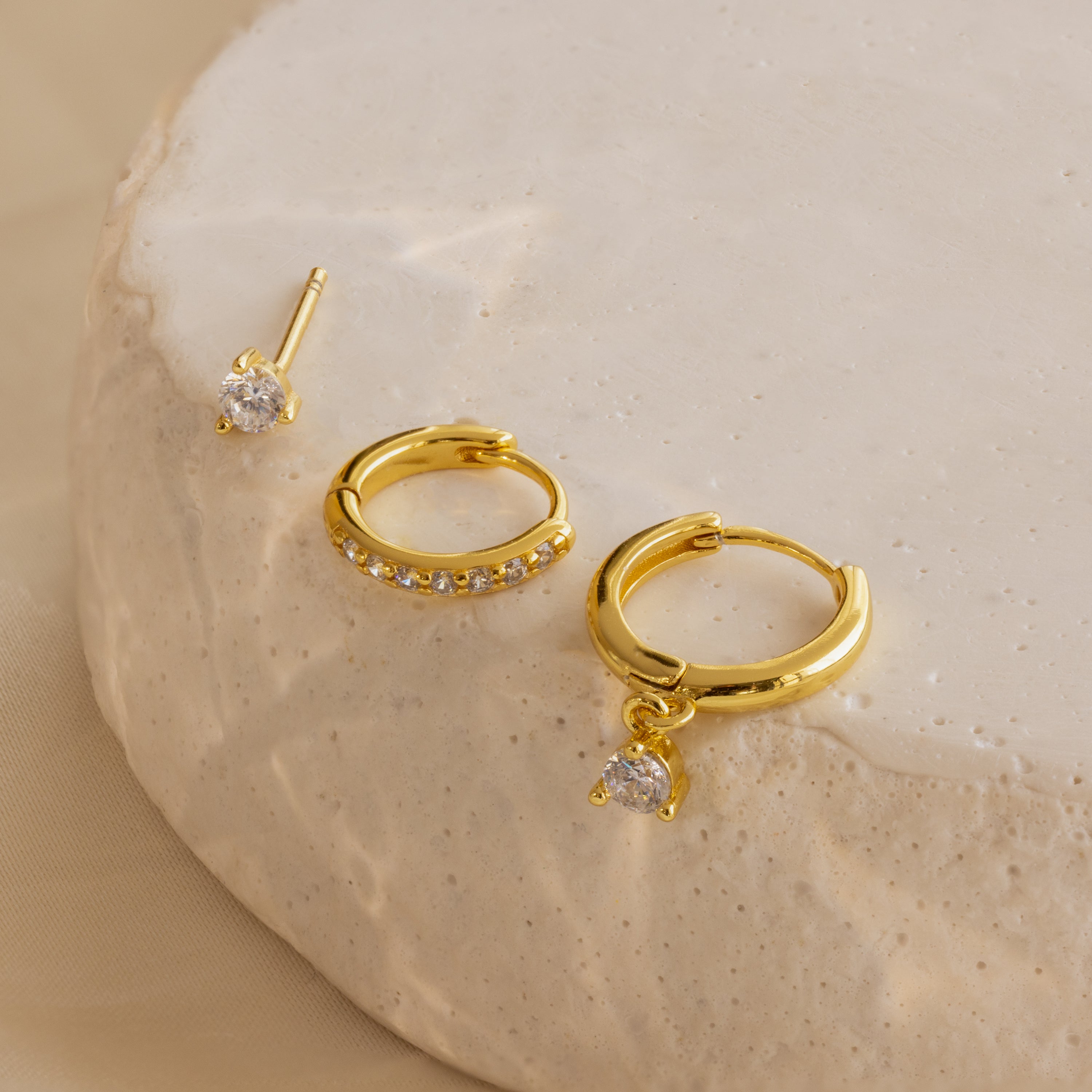 The Classic Diamond Earrings Set in 18K Gold features three pairs with clear gemstones, displayed on a textured beige surface—perfect for adding elegance to any bridal look.