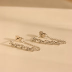 Diamond Chain Drop Earrings feature five small diamonds on each earring and are elegantly displayed against a cream-colored surface.