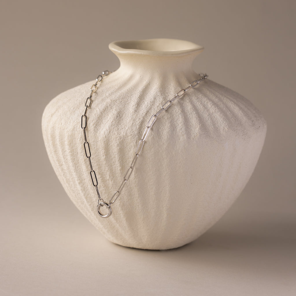 The Paperclip Clasp Necklace in White Gold is elegantly draped over a textured white ceramic vase against a neutral background.