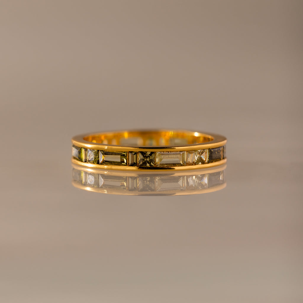The Peridot Inlay Eternity Ring features a geometric engraved gold band with a subtle peridot inlay, shown on a reflective surface against a neutral background.