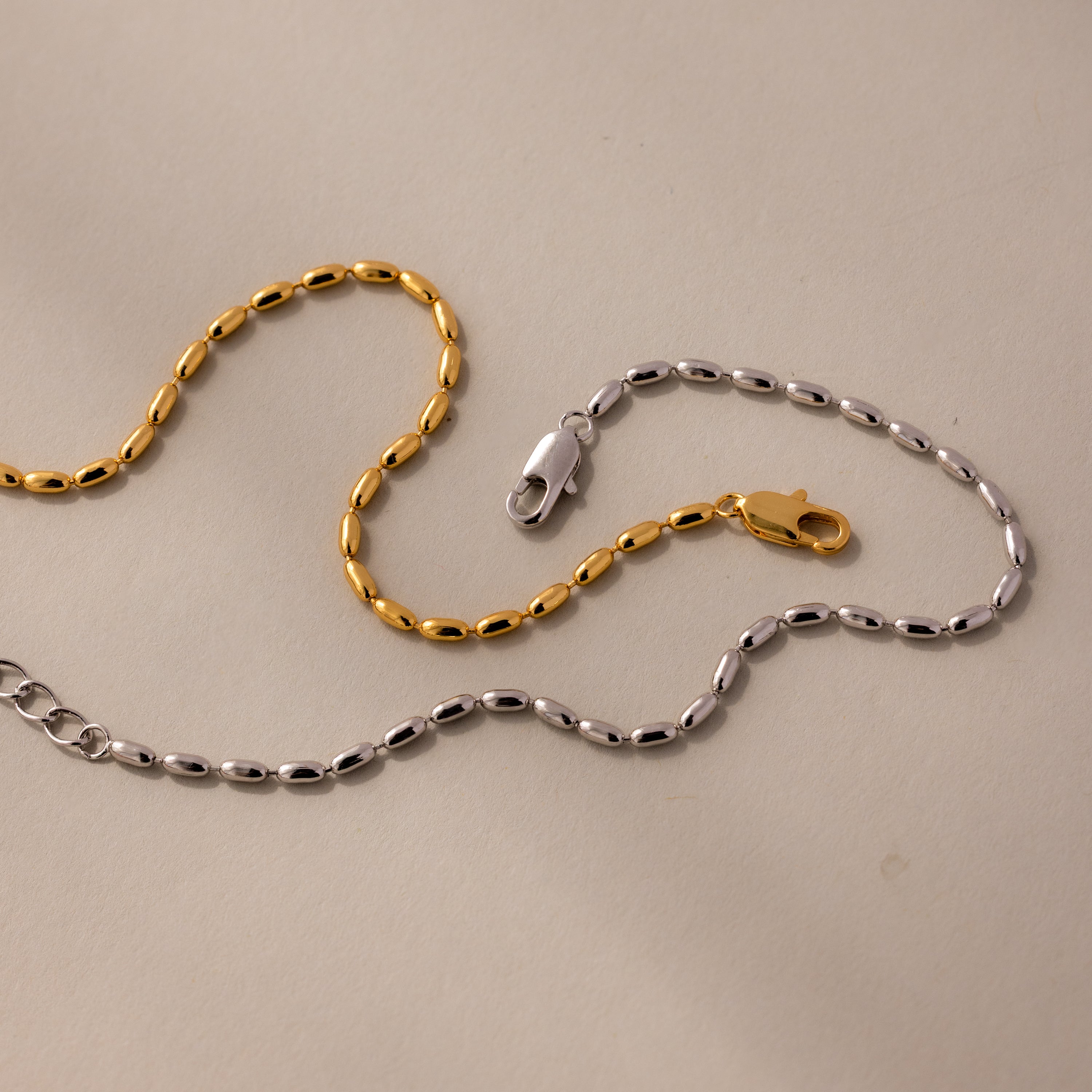 Gold and silver chain necklaces with oval links and clasps, paired with the Oval Beaded Bracelet in 18K Gold, highlight modern jewelry elegance against a neutral background.