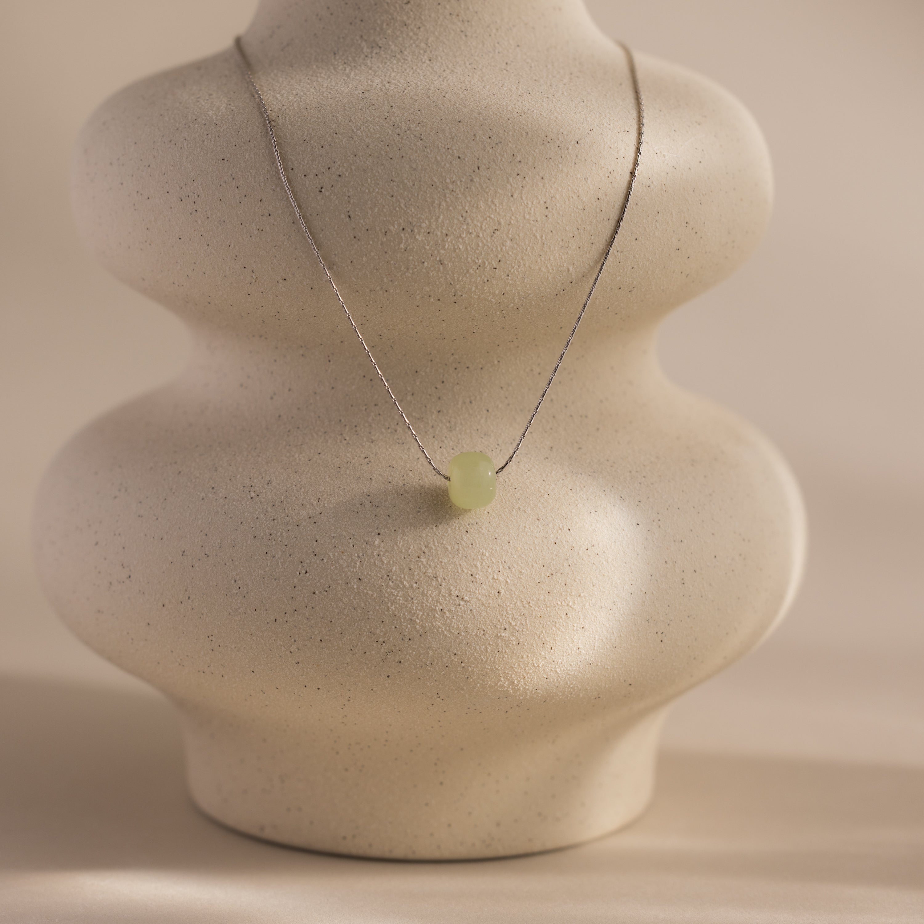 Delicate silver chain necklace featuring a single round green jade bead, displayed on a ceramic prop.