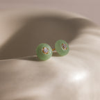 Close-up of round green jade stud earrings featuring a sparkling CZ diamond center and gold bezel setting, displayed on a ceramic dish.