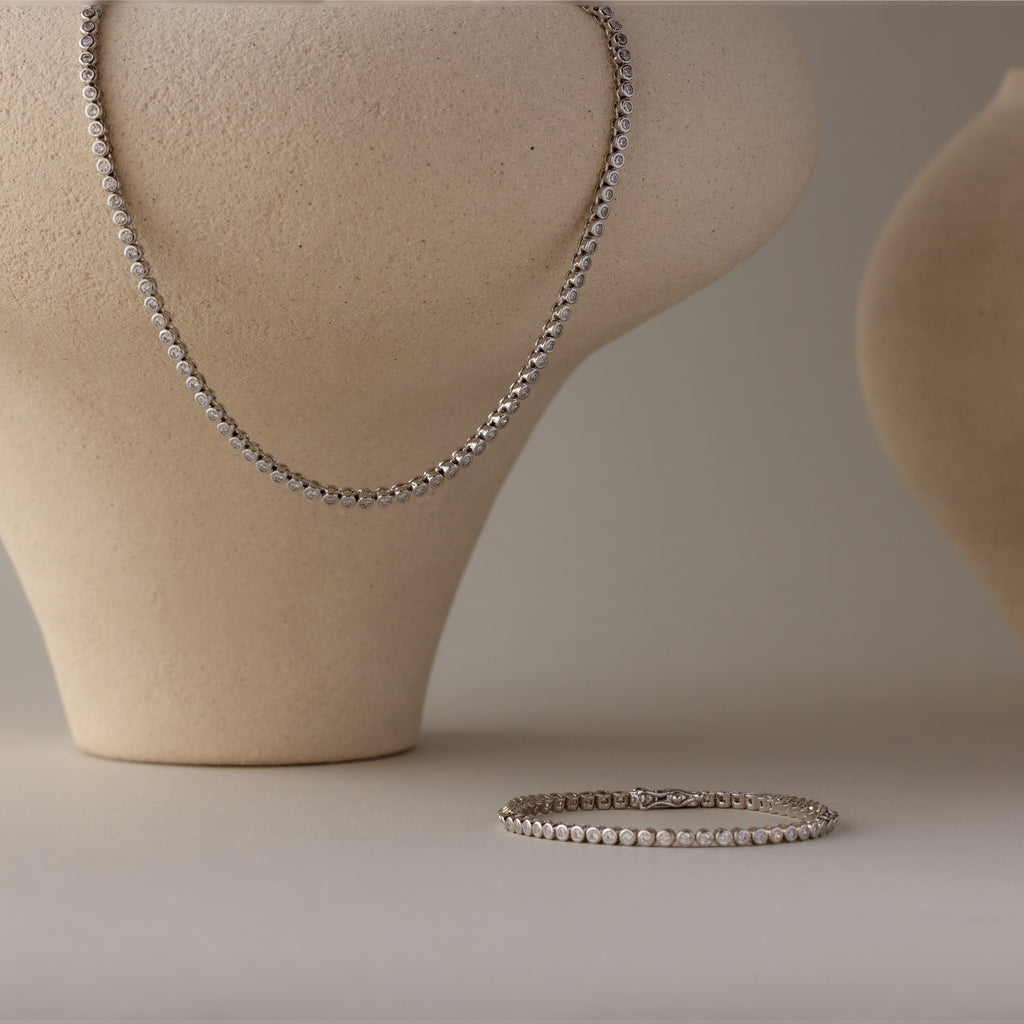 The Diamond Bezel Tennis Necklace & Bracelet Set in White Gold is elegantly displayed on neutral sculpted stands, making it a stunning anniversary gift against a simple background.