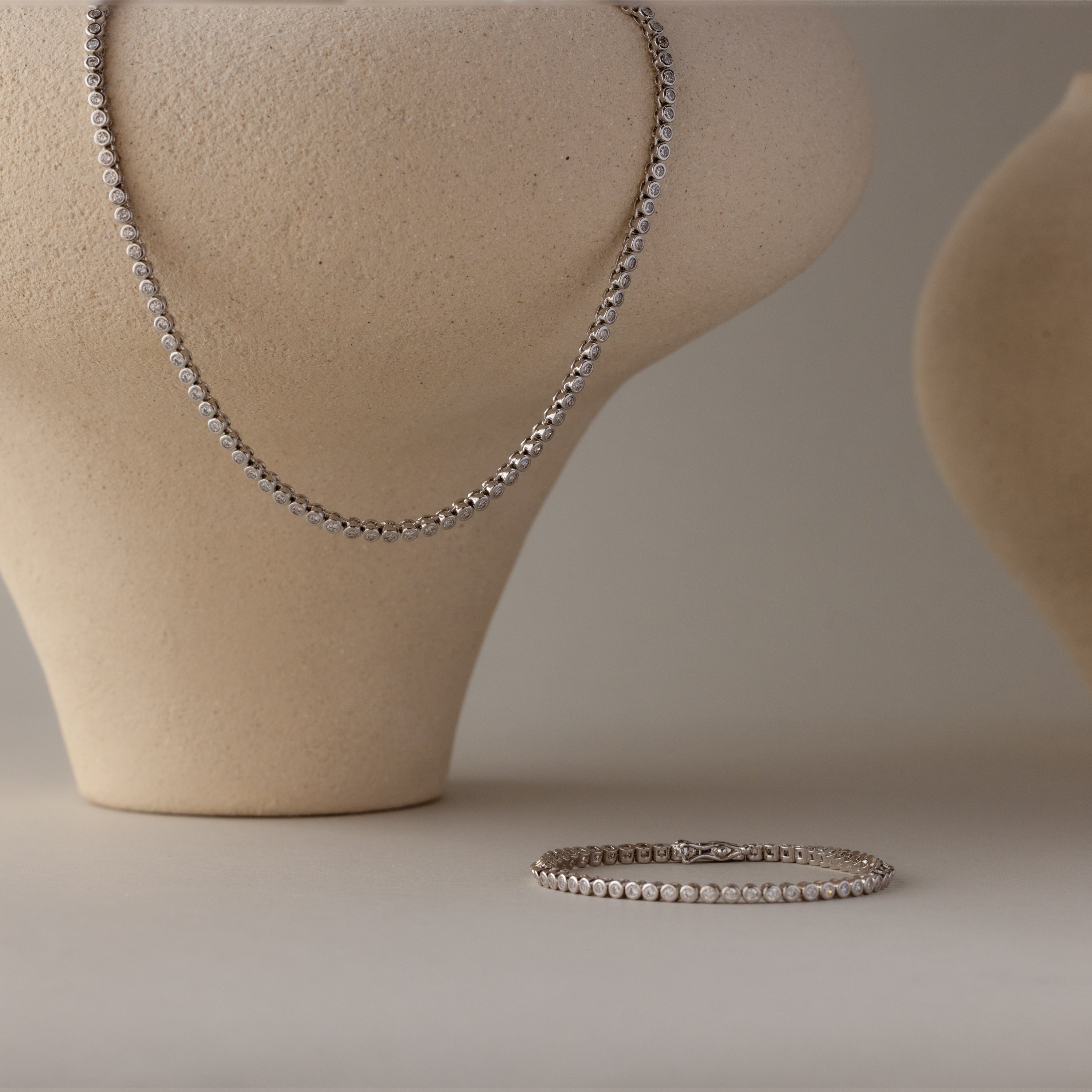 The Diamond Bezel Tennis Necklace & Bracelet Set in White Gold is elegantly displayed on neutral sculpted stands, making it a stunning anniversary gift against a simple background.