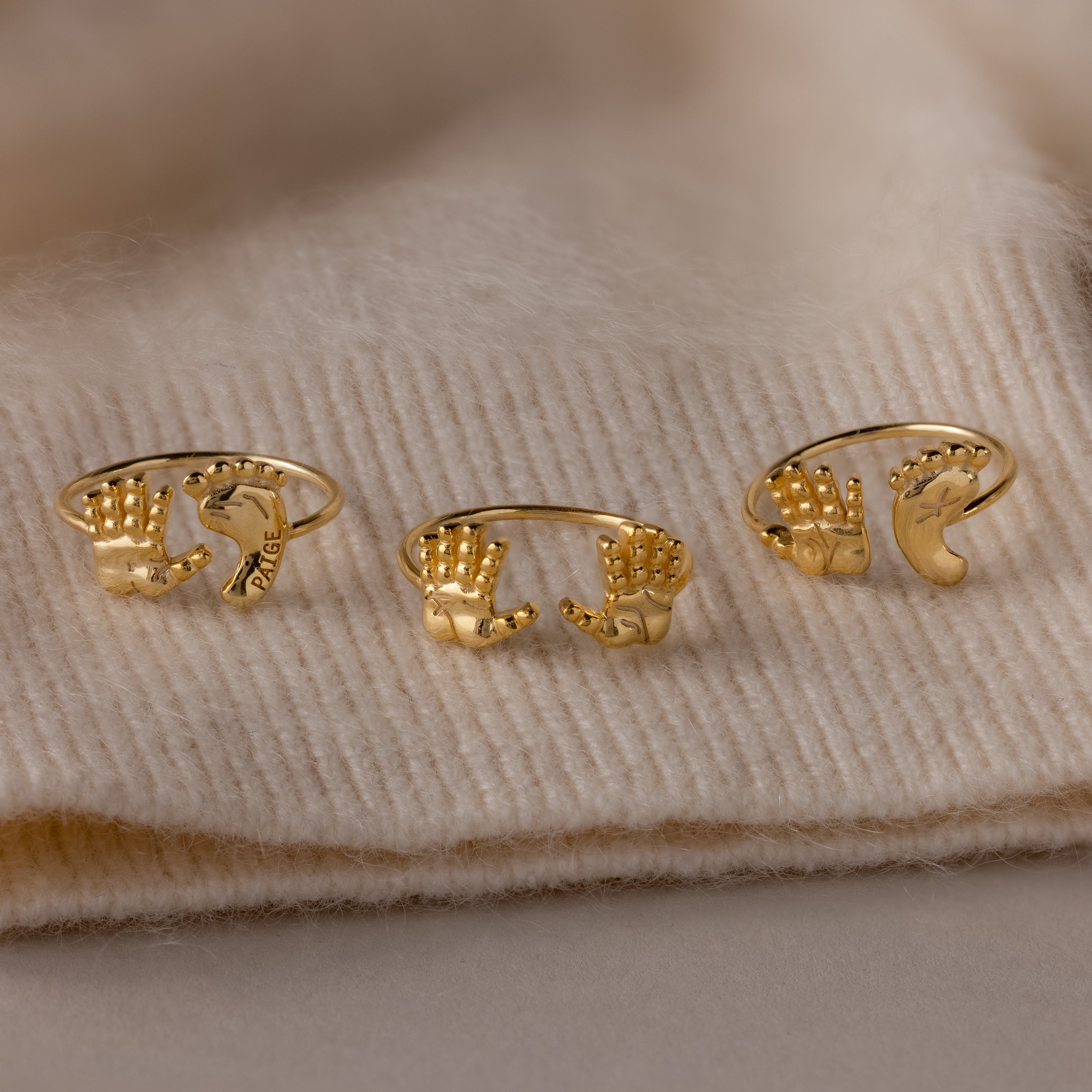 Three Hand & Footprint Duo Name Rings in 18K Gold, featuring unique hand-shaped designs, are beautifully displayed on a beige knitted fabric background—an ideal keepsake gift for new parents.