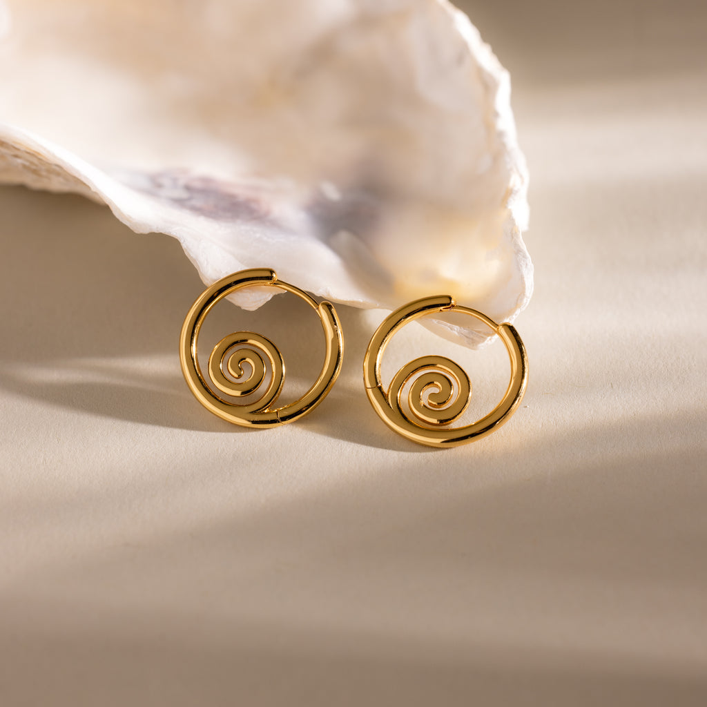 Spiral Swirl Hoops in 18K Gold displayed before a white seashell on beige, perfect beachy earrings to complete your next boho vacation jewelry collection.