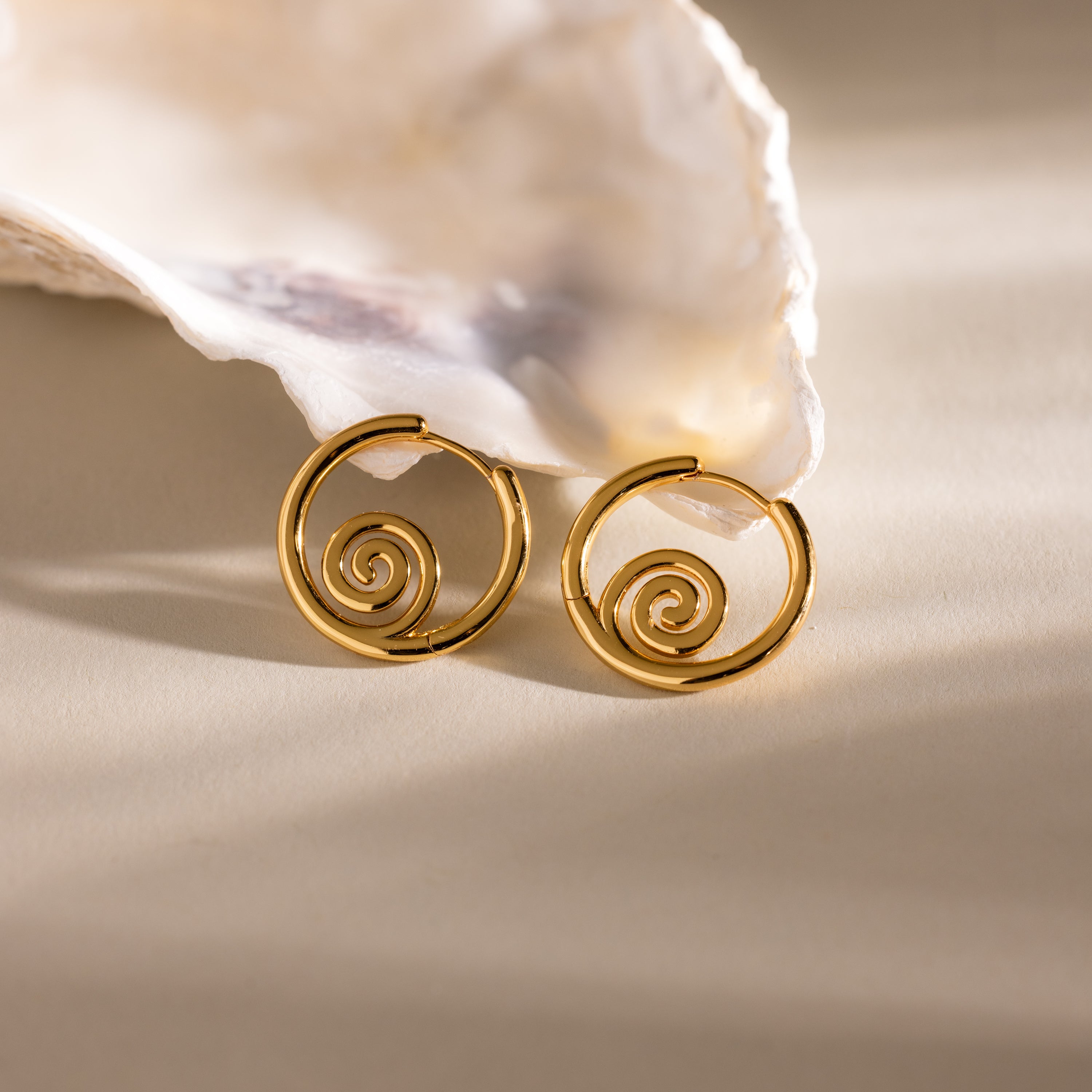 Spiral Swirl Hoops in 18K Gold displayed before a white seashell on beige, perfect beachy earrings to complete your next boho vacation jewelry collection.