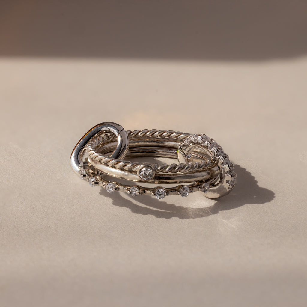 A stack of silver rings with twisted and smooth bands, some set with clear gemstones, rests on a light surface—ideal for customizing or pairing with the Multipurpose Pave Ring Lock Set in White Gold.