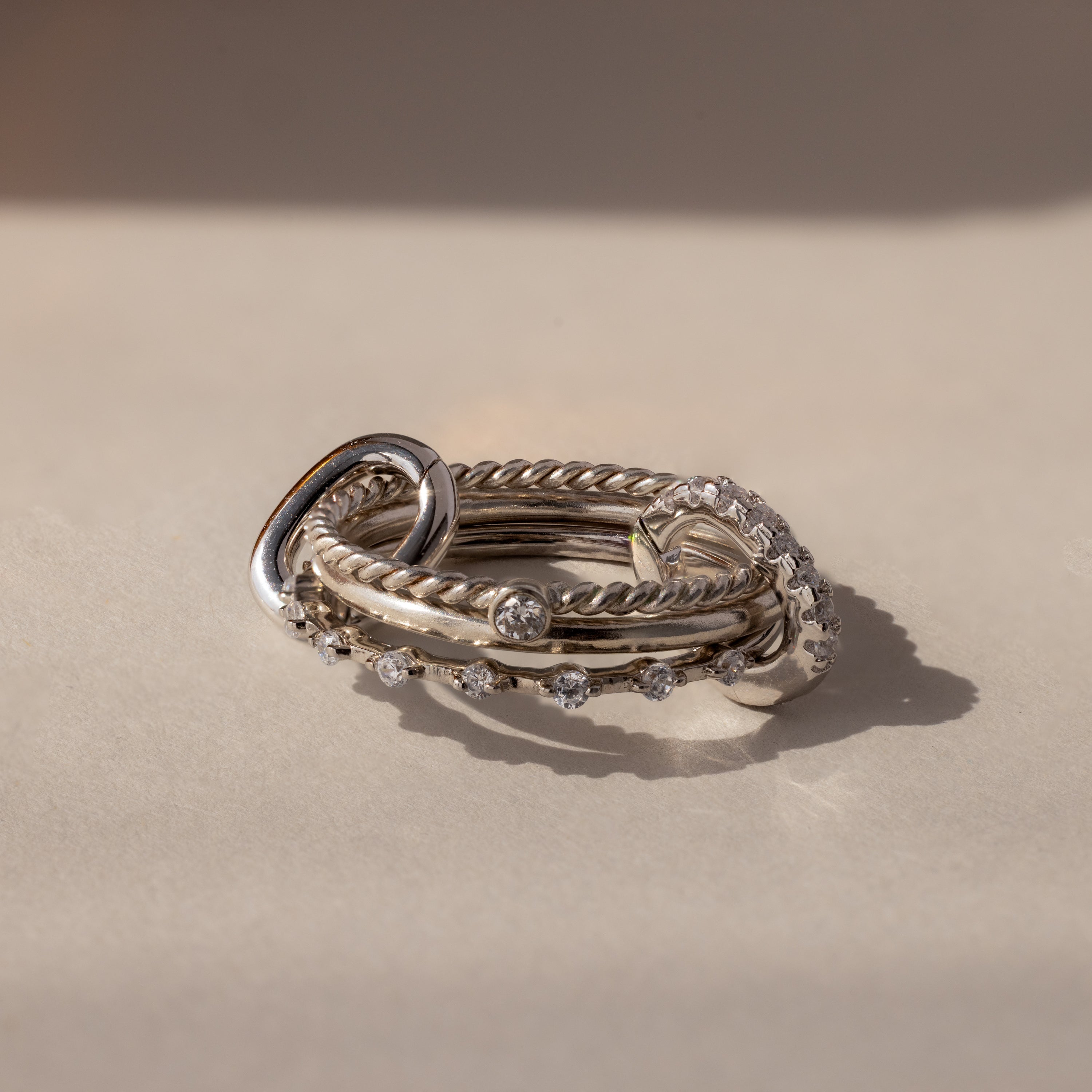 A stack of silver rings with twisted and smooth bands, some set with clear gemstones, rests on a light surface—ideal for customizing or pairing with the Multipurpose Pave Ring Lock Set in White Gold.