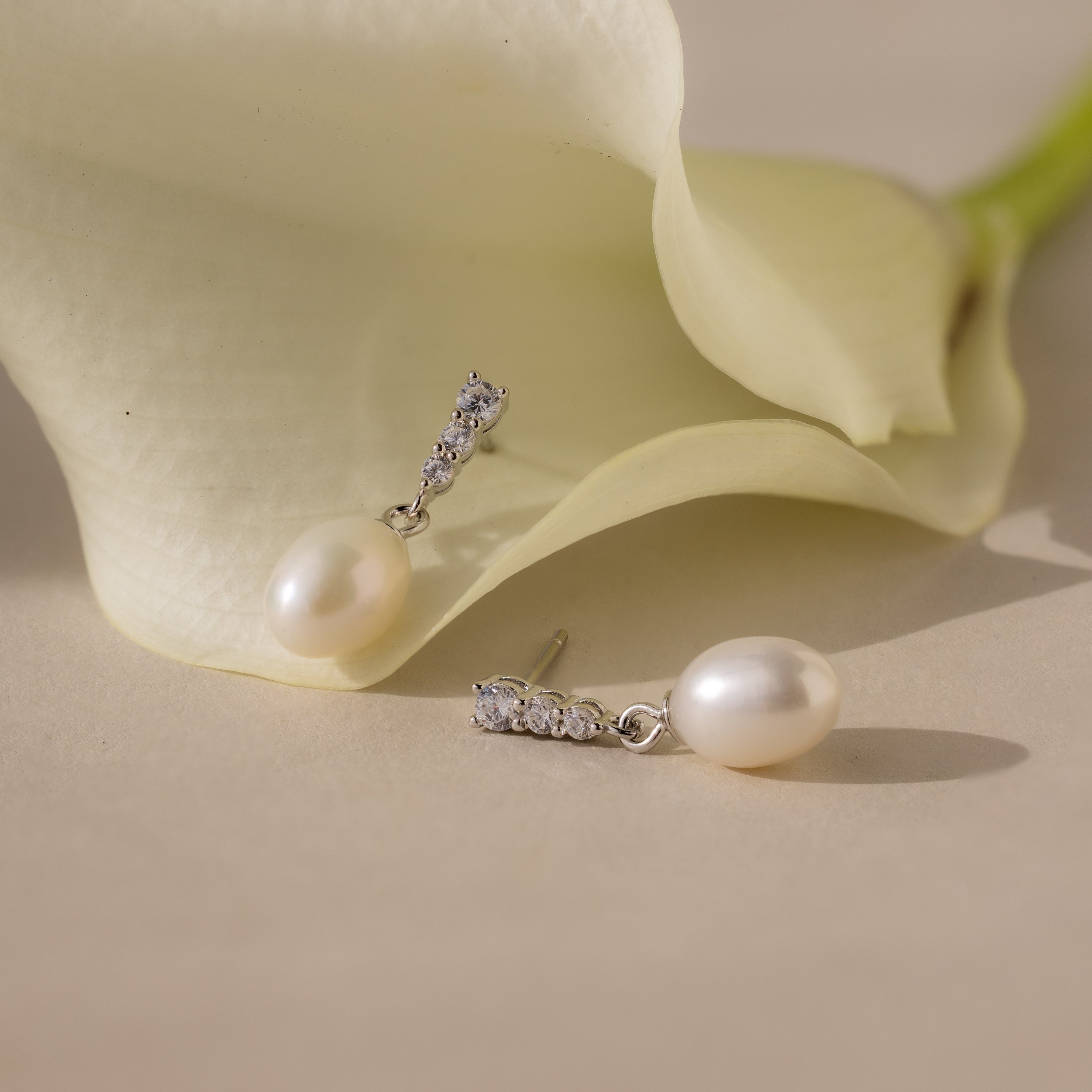 The Diamond Pearl Droplet Earrings in Sterling Silver are displayed beside a white calla lily on a beige surface.