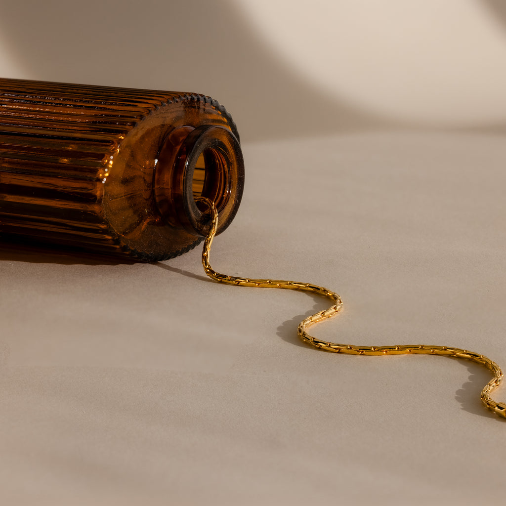 The Bold Link Chain Necklace in 18K Gold spills from a brown glass bottle lying on its side on a beige surface.