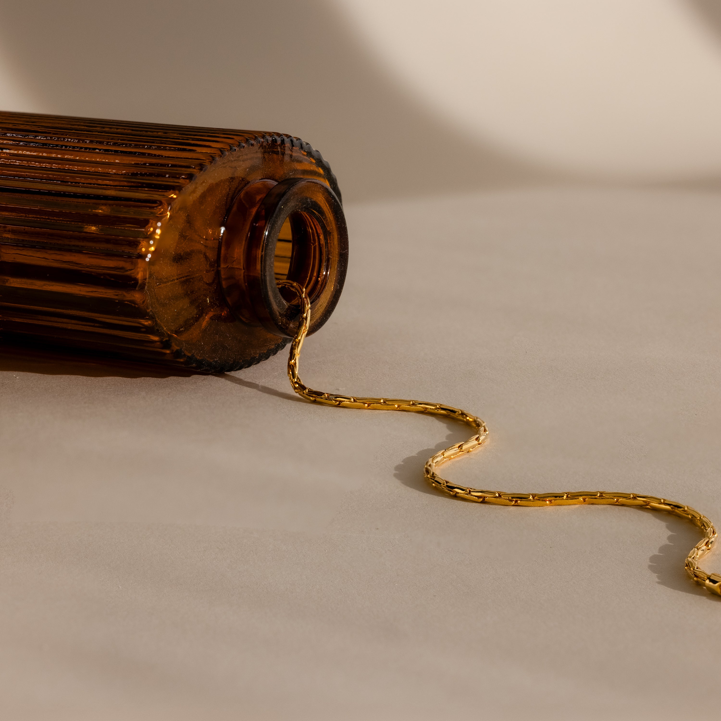 The Bold Link Chain Necklace in 18K Gold spills from a brown glass bottle lying on its side on a beige surface.