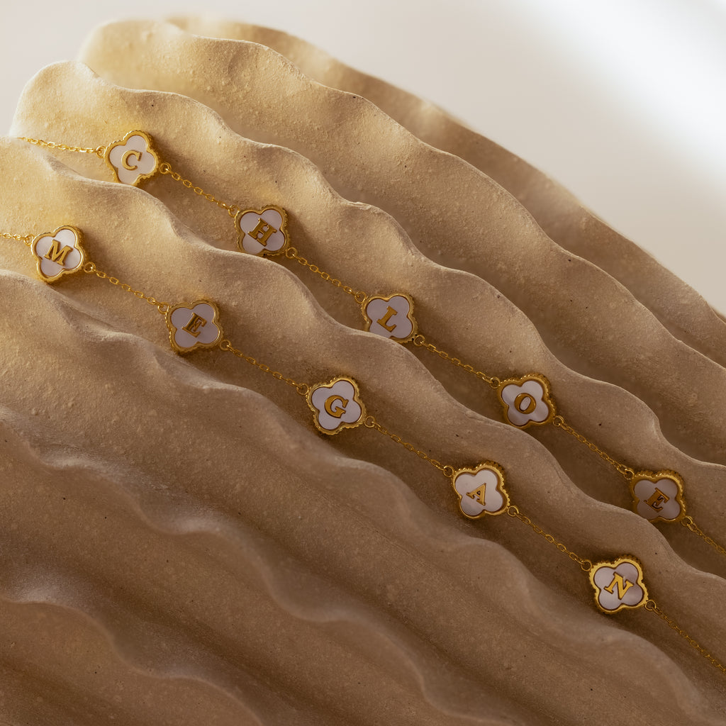 The Pearl Clover Letter Bracelet in 18K Gold is shown on wavy beige fabric, with its white floral charm and gold details creating an elegant, mother of pearl-inspired look.