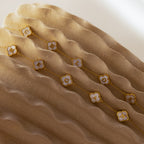 The Pearl Clover Letter Bracelet in 18K Gold is shown on wavy beige fabric, with its white floral charm and gold details creating an elegant, mother of pearl-inspired look.