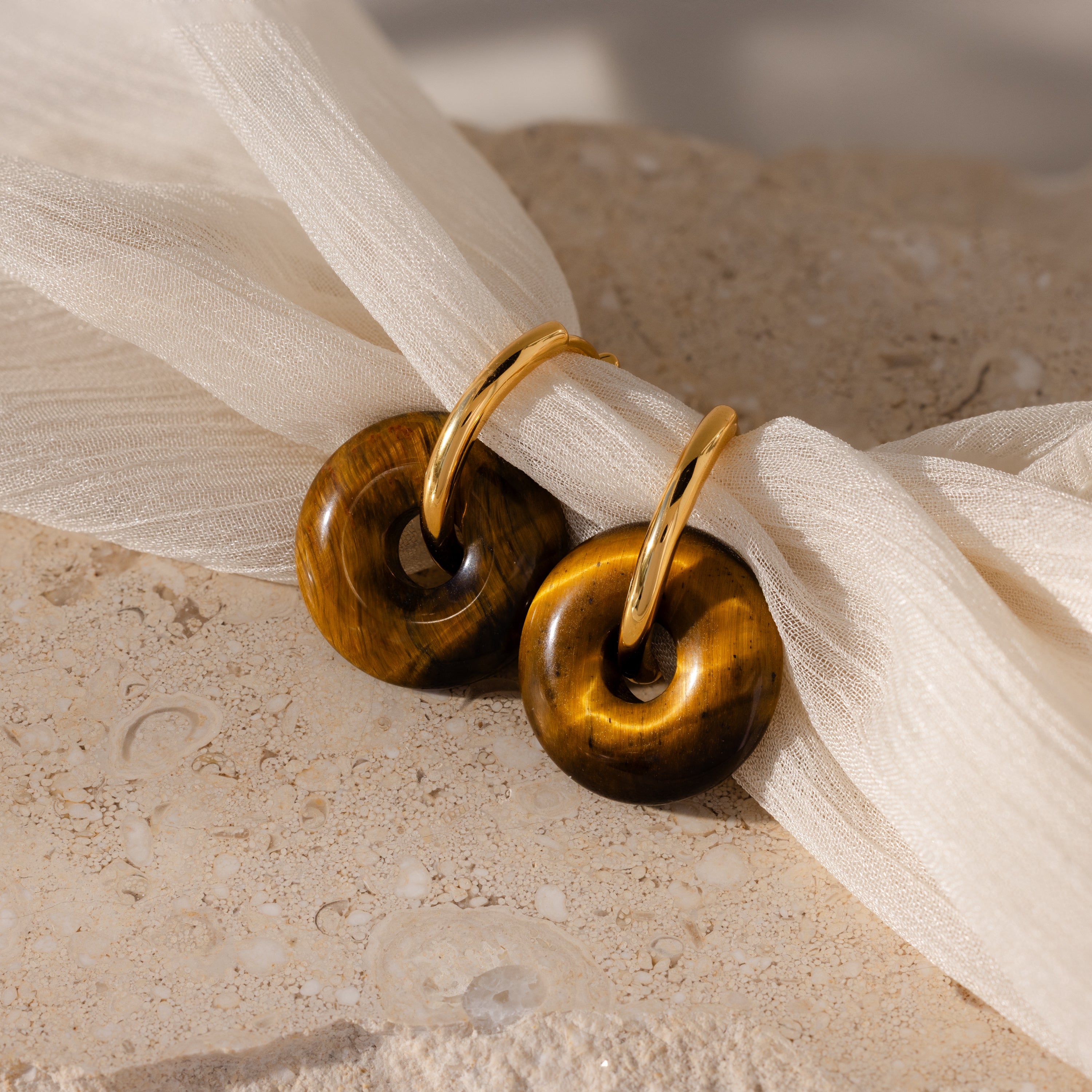 Tiger's Eye Duo Hoops in 18K Gold, featuring round Tiger's Eye stone pendants, rest on a textured beige surface and soft fabric, bringing bold jewelry elegance.