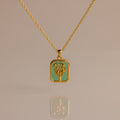 The Emerald Birth Flower Charm Necklace features a rectangular green pendant with a floral design, showcased on a neutral background for an elegant and timeless jewelry statement.