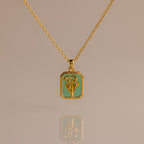 The Emerald Birth Flower Charm Necklace features a rectangular green pendant with a floral design, showcased on a neutral background for an elegant and timeless jewelry statement.