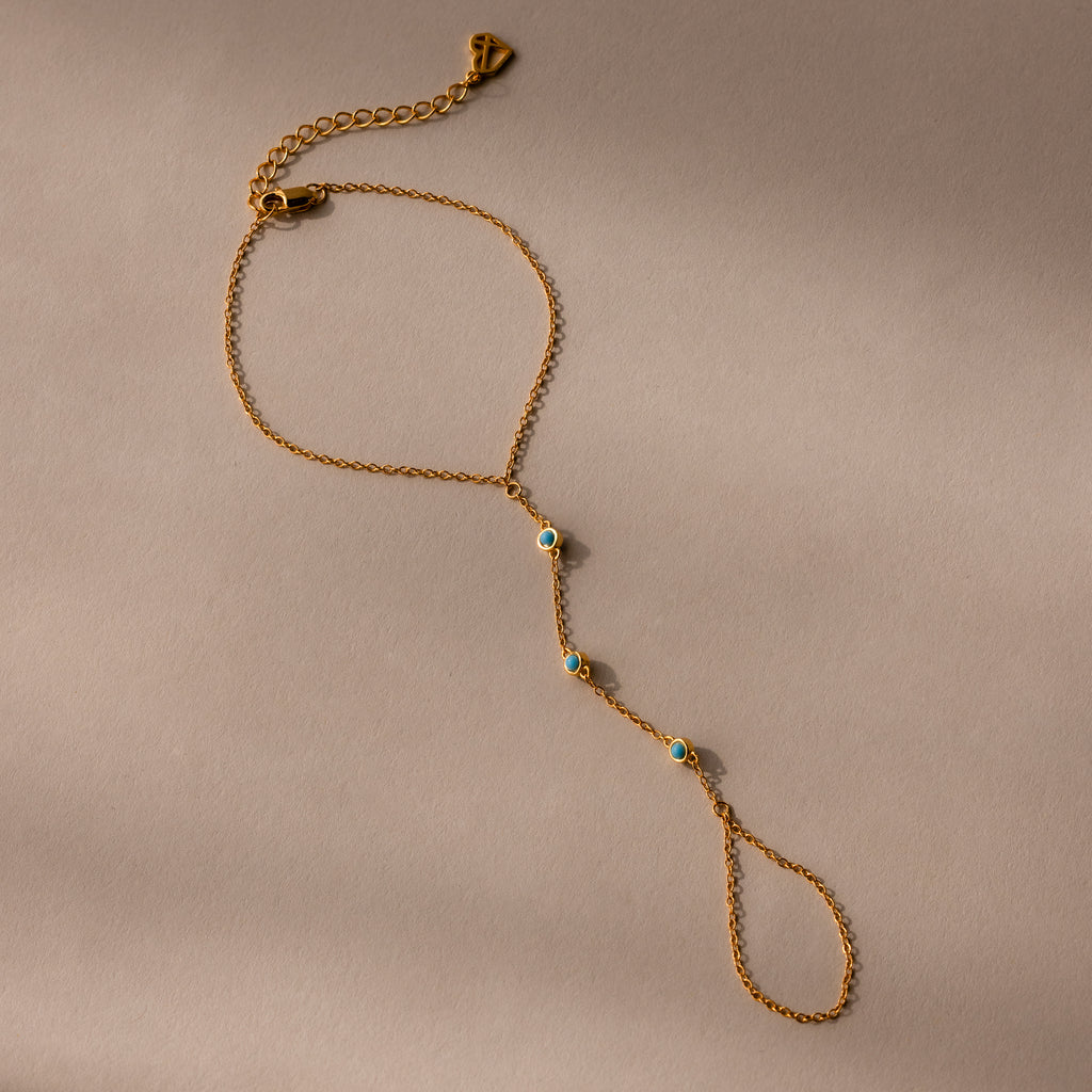The Turquoise Hand Chain Bracelet in 18K Gold features a delicate design with three small blue and white stones, shown against a beige background.