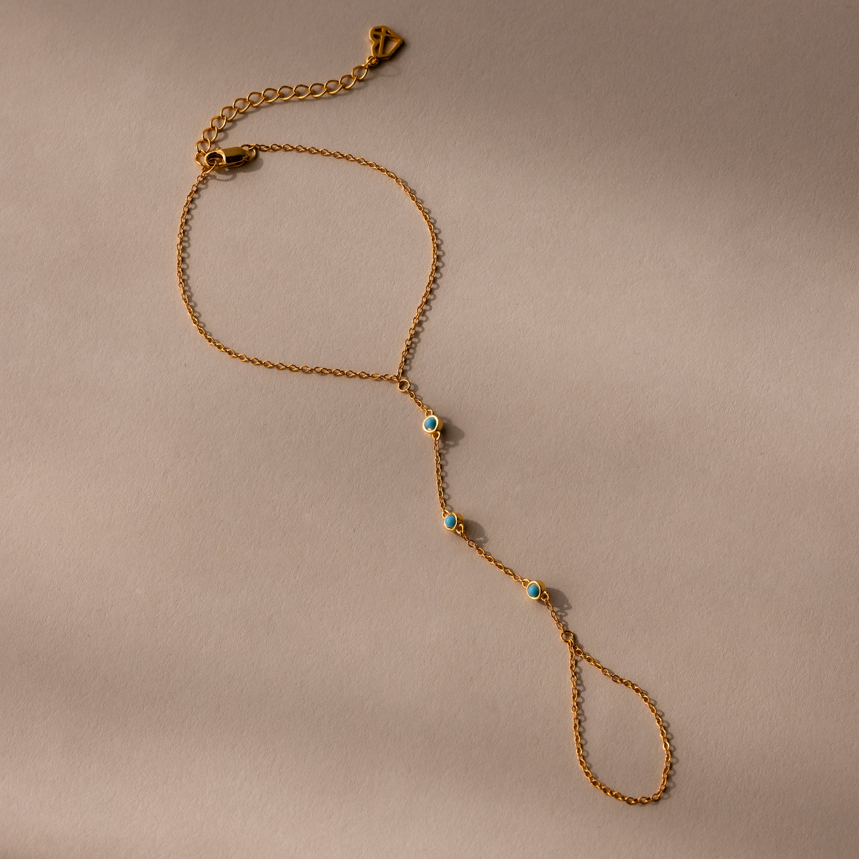 The Turquoise Hand Chain Bracelet in 18K Gold features a delicate design with three small blue and white stones, shown against a beige background.