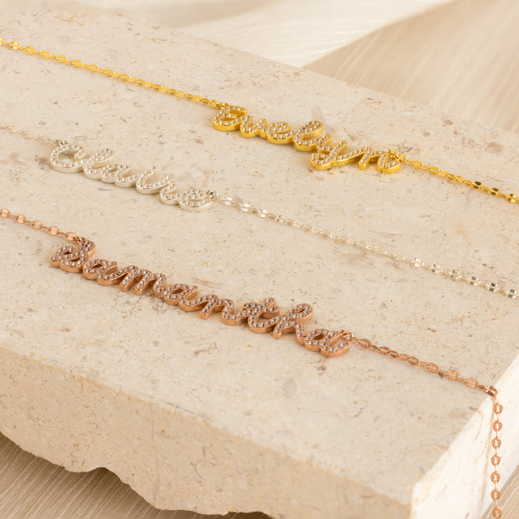 Three Pave Dream Script Name Necklaces with the names evelyn, claire, and samantha in gold, silver, and rose gold rest on a stone slab—a stylish update to the classic custom necklace.