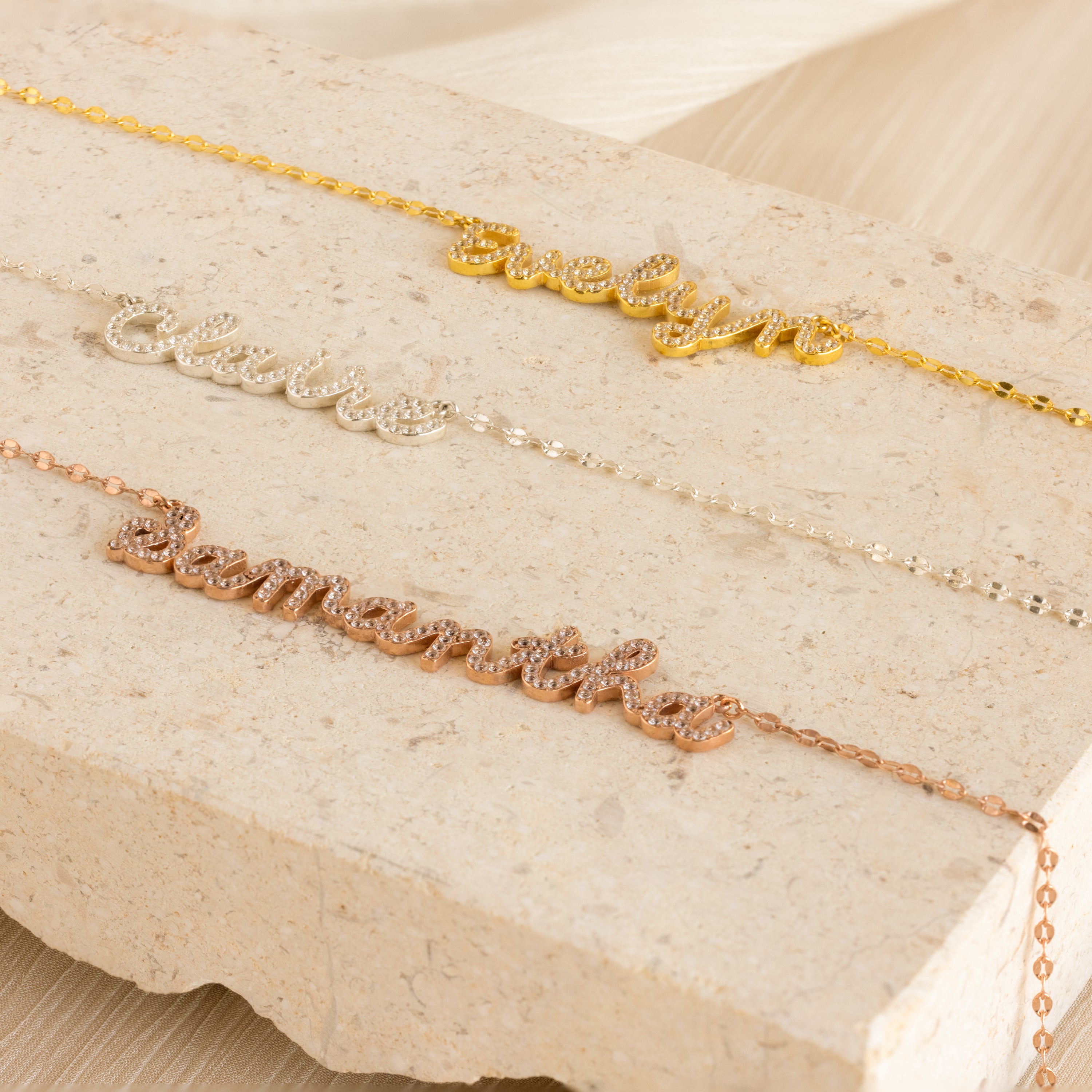 Three Pave Dream Script Name Necklaces with the names evelyn, claire, and samantha in gold, silver, and rose gold rest on a stone slab—a stylish update to the classic custom necklace.