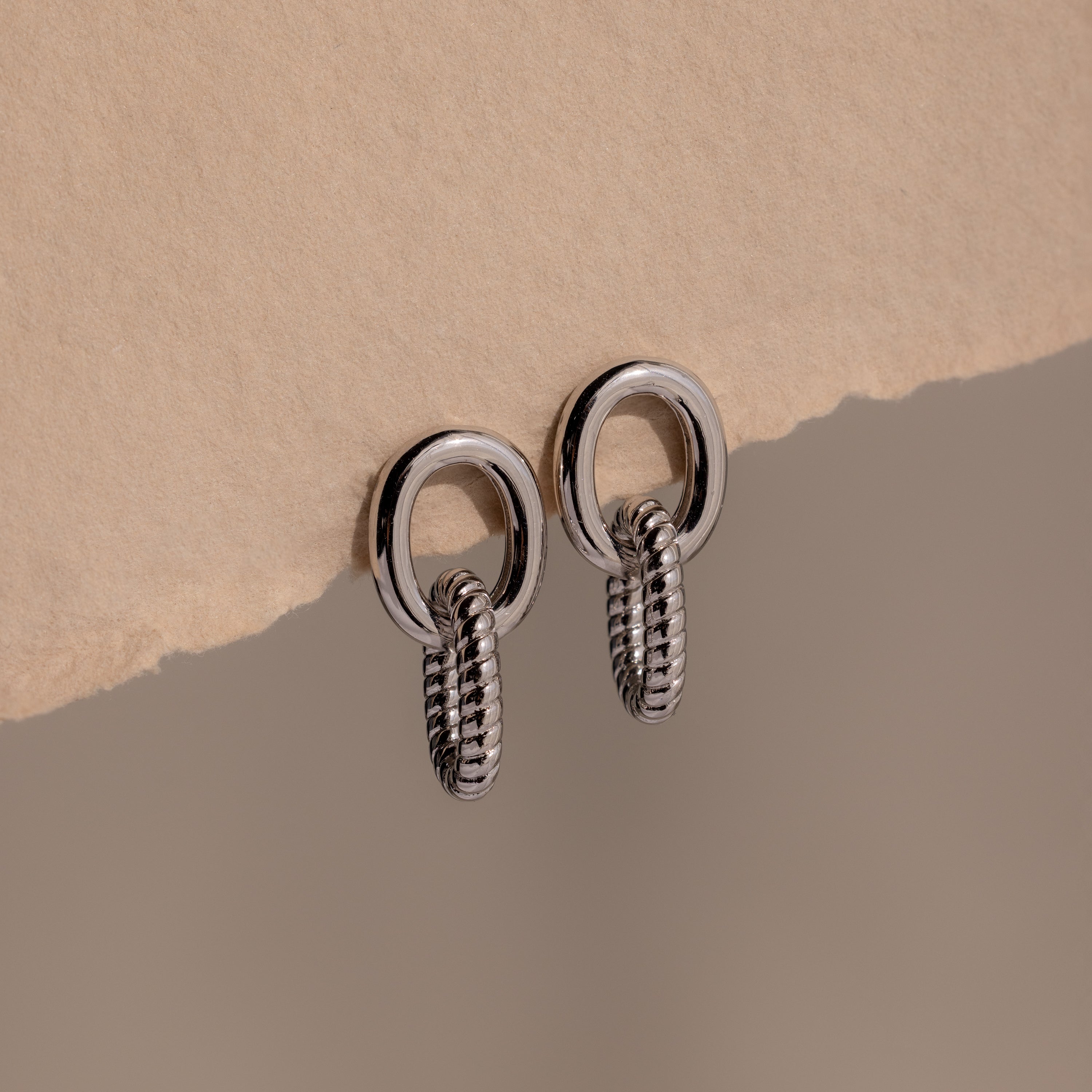 Twisted Link Earrings in White Gold featuring textured chain links are showcased on a torn beige paper background—a perfect blend of modern design and classic jewelry style.