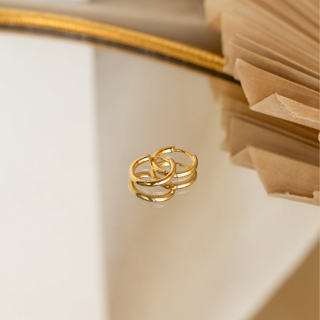 Small Bold Hoops displayed on a reflective surface with beige decorative paper and a gold-edged mirror, highlighting their minimalist design—perfect for everyday wear or pairing with other earrings.